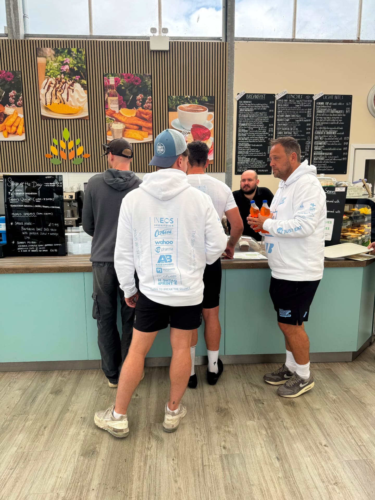 Cafe stop, mid-stage Mitch Mottram and riders in The Longest Round sponsor hoodies queueing at a roadside cafe counter during a rest stop