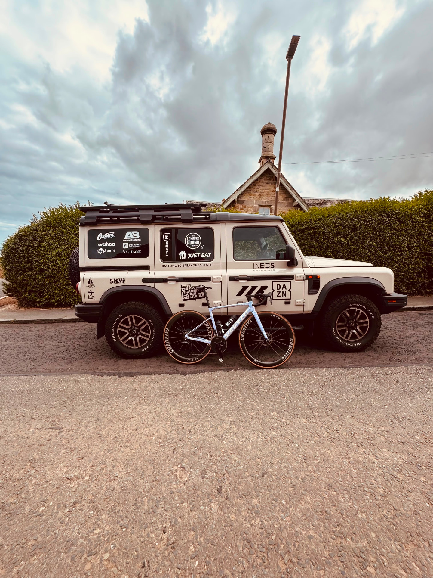 Bike and support vehicle at first light Road bike leaning against the Longest Round support vehicle Mitch Mottram crewed on, branded INEOS Grenadier support vehicle under heavy Scottish cloud