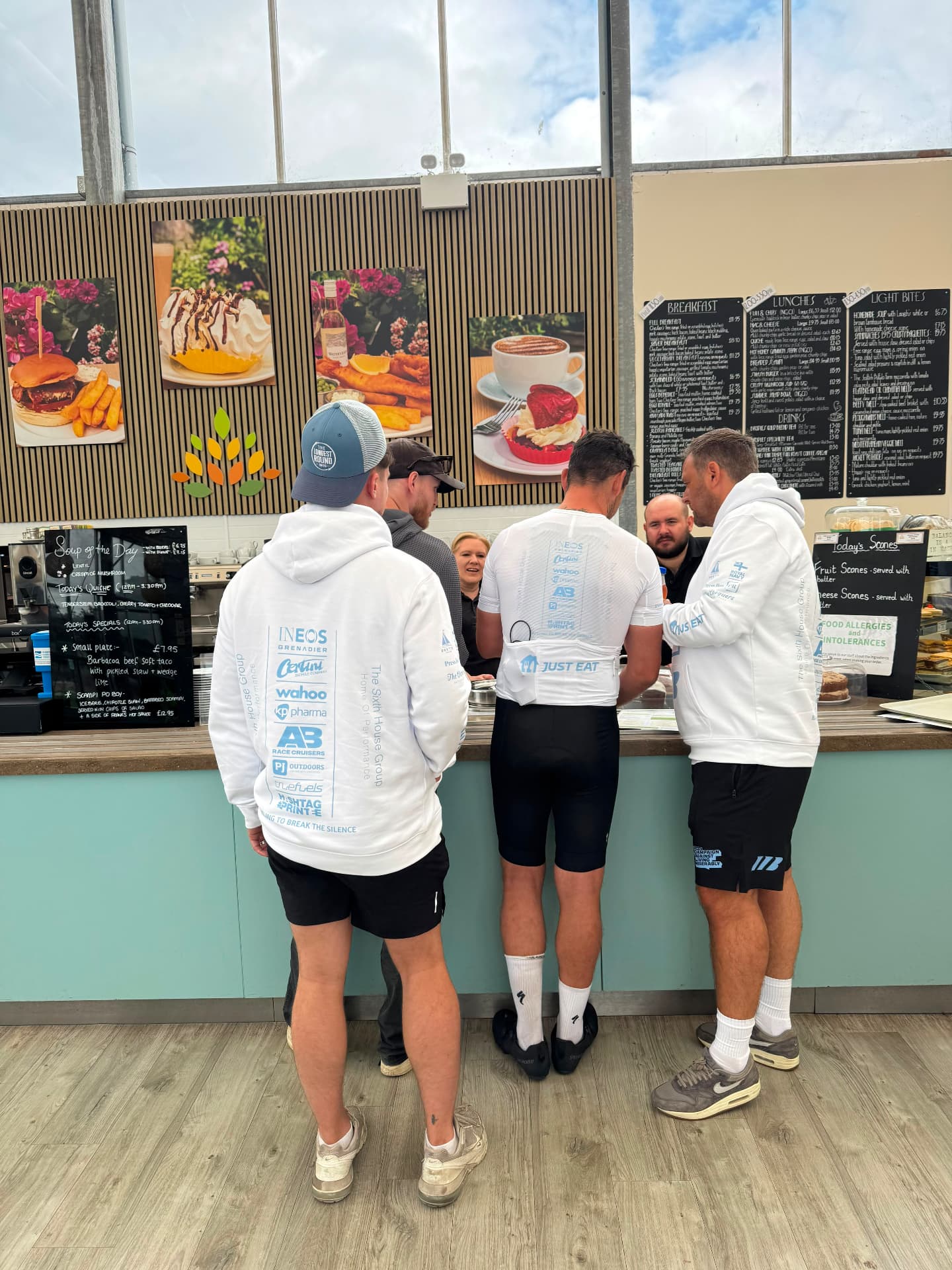 Mitch Mottram and cyclists in kit refuelling with drinks at a cafe counter between stages of The Longest Round
